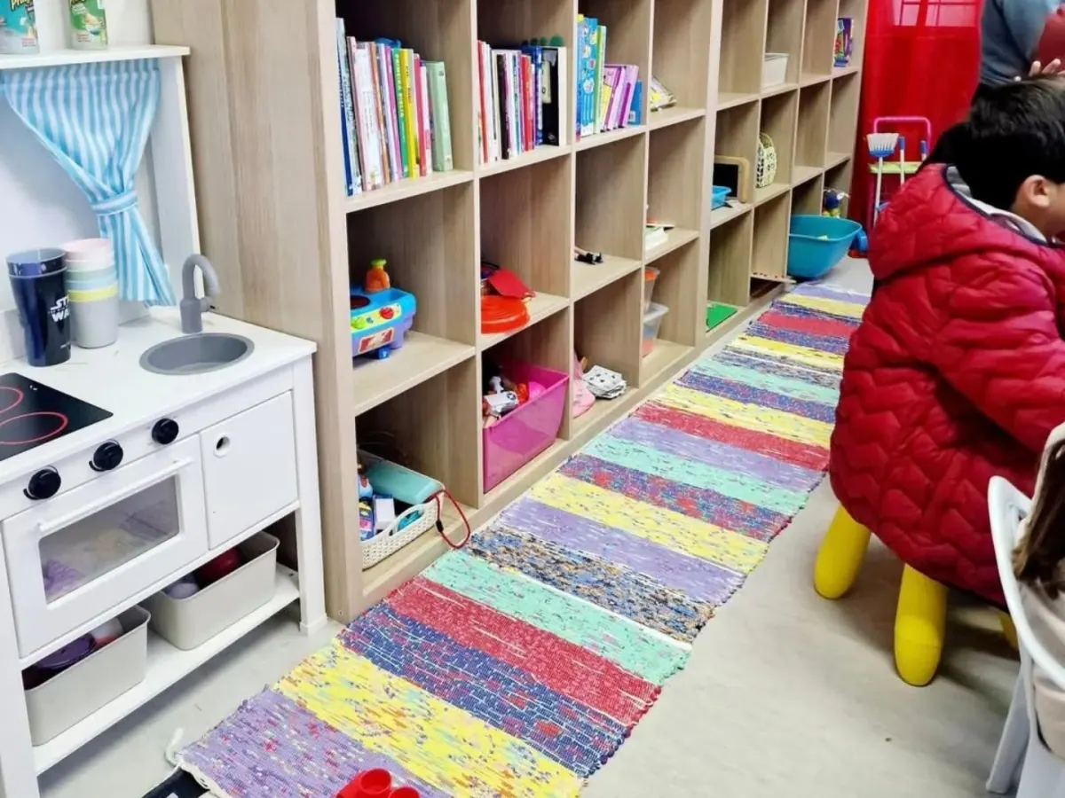 Sala de ATL com estantes de livros e brinquedos, cozinha de brincar e tapete de riscas coloridas